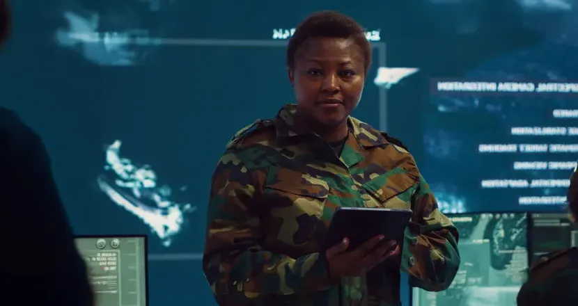 Military service member in camouflage uniform holding a tablet in a high-tech operations center with digital surveillance screens in the background.